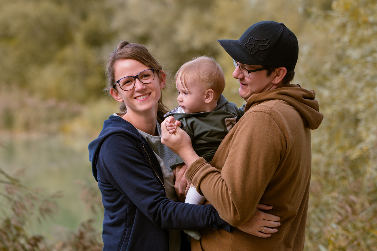 Junges Elternpaar steht eng beisammen und hält ihr Baby im Arm; die Mutter lächelt warm in die Kamera, während Vater und Kind einander zugewandt sind, umgeben von natürlicher Landschaft.